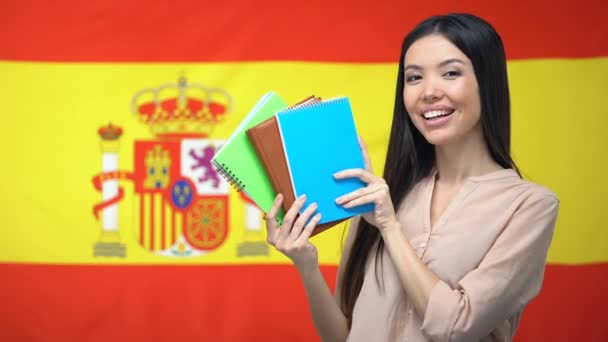 Азиатская девушка показывает тетради на фоне испанского флага, изучает язык — стоковое видео