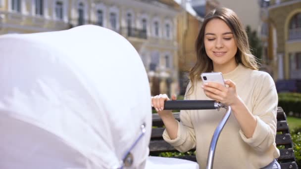 Joyful young mother reading internet forum on smartphone swinging baby carriage — Stock Video