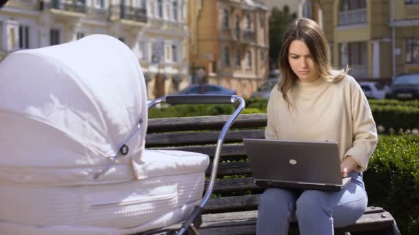 Sibuk perempuan muda mencoba untuk bekerja laptop dan mengayunkan bayi yang baru lahir di kereta bayi, perawatan anak — Stok Video