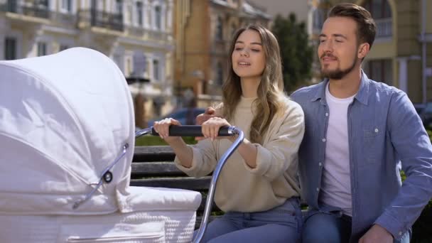 Joyful jovem casal admirando bebê em carruagem sentado no parque de banco, amor familiar — Vídeo de Stock