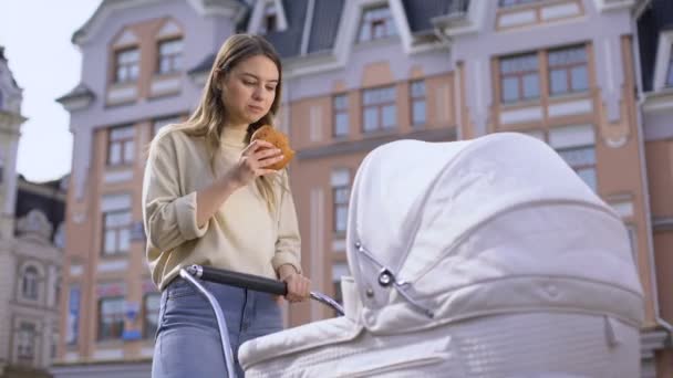 紧张忙碌的妈妈摆动婴儿在婴儿车和咀嚼包子，有小吃在公园 — 图库视频影像
