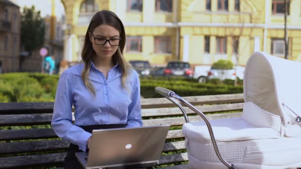 Carreira laptop de trabalho feminino e bebê balançando no carrinho sentado no banco do parque — Vídeo de Stock