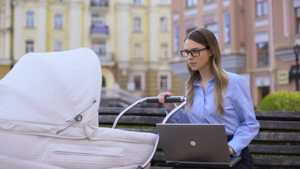 Multitarea madre balanceo bebé carro y portátil de trabajo sentado en el banco — Vídeos de Stock