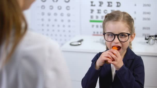 Счастливый ребенок в очках ест морковь по рекомендации врача, бета-каротин — стоковое видео