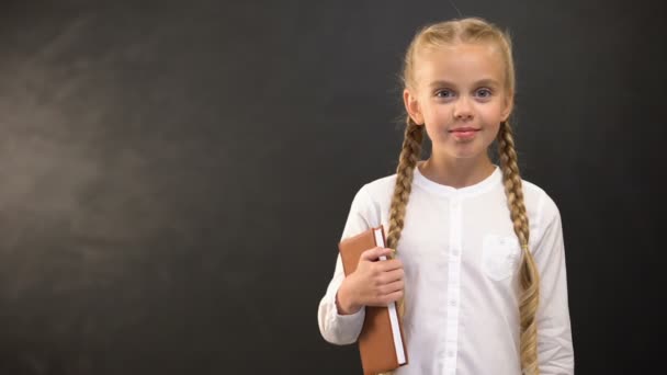 Schoolmeisje met boek en stukje krijt glimlachend op camera tegen Blackboard — Stockvideo