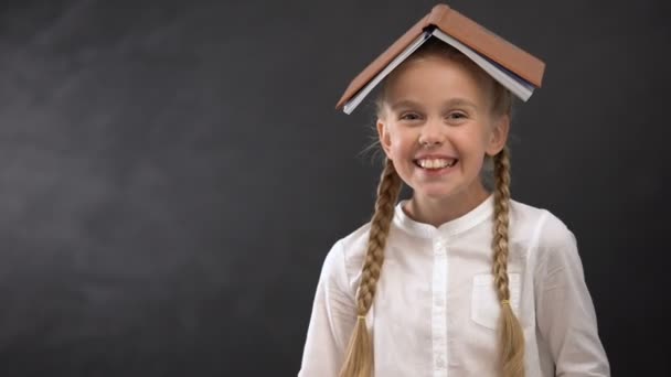 Возбужденная ученица с книгой на голове, улыбающаяся в камеру, концепция образования — стоковое видео
