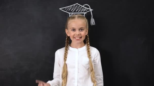 Happy schoolgirl points finger at academic cap painted on blackboard background — Stock Video