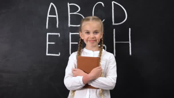 Livre de câlins écolière souriant, alphabet écrit sur tableau noir derrière, système — Video