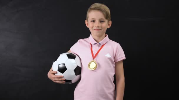 Niño con pelota y medalla de pie cerca de pizarra, ganador de la competición de fútbol — Vídeos de Stock