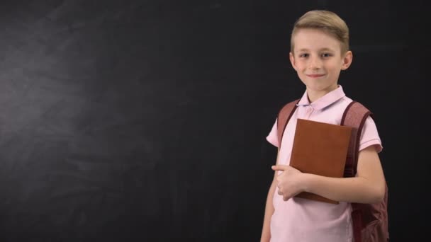 Escolar diligente sosteniendo libro de texto, de pie cerca de pizarra, sistema educativo — Vídeos de Stock