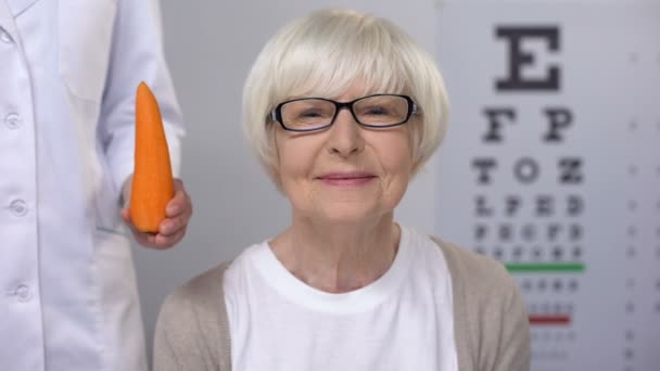 Oculist yaşlı bayan gülümseyen havuç öneren, görme için doğal vitaminler — Stok video