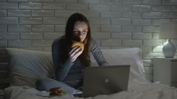 Femeie tânără care mănâncă hamburger în timp ce navighează pe web noaptea pe pat, junk food — Videoclip de stoc
