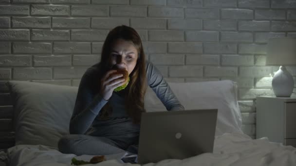 おいしい甘やかされたハンバーガーを噛む女性、ベッドで夜にジャンクフードの食事を持っている — ストック動画