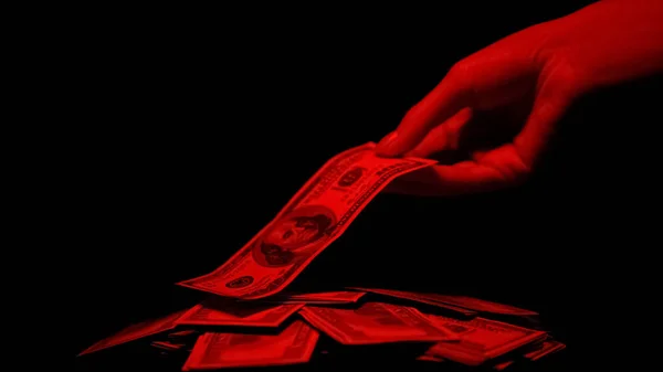 手从红光照亮的堆里拿美元 血淋淋的金钱犯罪奖励 — 图库照片