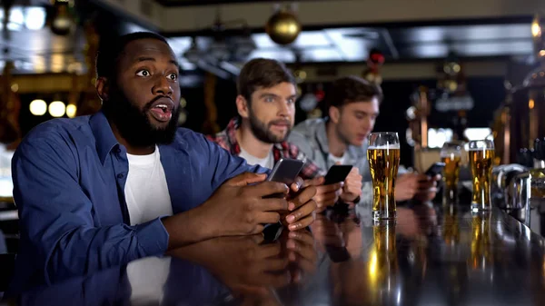 アフリカ系アメリカ人の男性がスマートフォンで賭けをチェックし バーで試合を見て アプリ — ストック写真