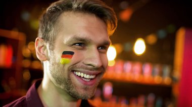 Yanağında bayrak olan neşeli Alman futbol taraftarı, gol atan takım için mutlu