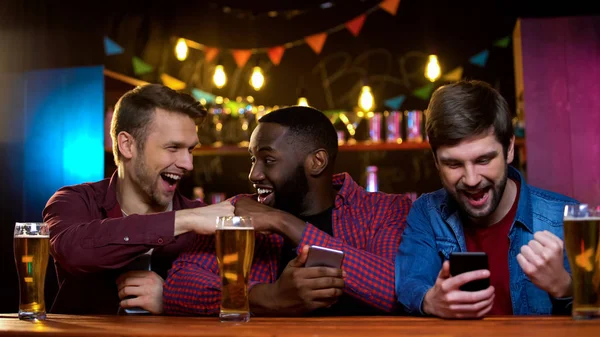 Male friends giving fist-bump sitting in beer pub, rejoicing online results