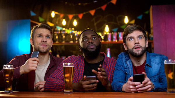 Multiethnic male friends watching football match in pub, upset with team losing