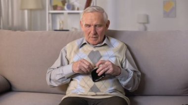 Üzgün kıdemli adam kanepede oturan ve kamera, yoksulluk boş cüzdan gösteren