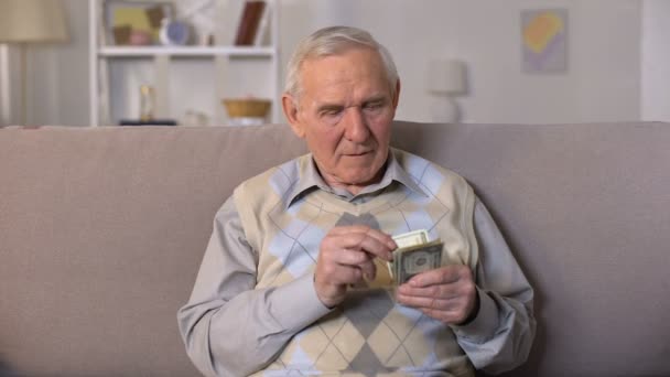 Depressed old man sitting on sofa, holding dollars banknotes, social insecurity — Stock Video