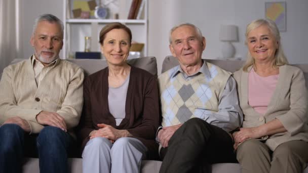 Веселые старшие друзья сидят на диване и улыбаются в камеру, счастливого выхода на пенсию — стоковое видео