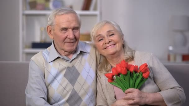 Пожилая пара обнимается дома, женщина держит тюльпаны, подарок на годовщину — стоковое видео