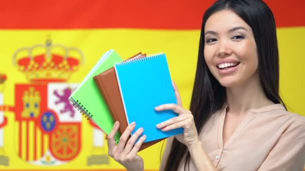 Mujer sonriente mostrando cuadernos a cámara, fondo bandera de España, estudiar en el extranjero — Vídeo de stock