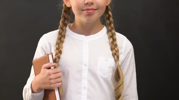 Маленька школярка стоїть з книгою і крейдою проти дошки, освіта — стокове відео
