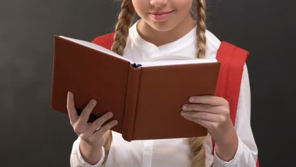 Alegre colegiala leyendo libro y sonriendo a la cámara contra pizarra — Vídeos de Stock