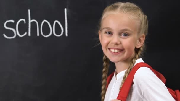 Petite fille avec sac à dos souriant à la caméra, mot de l'école écrit sur le tableau noir — Video