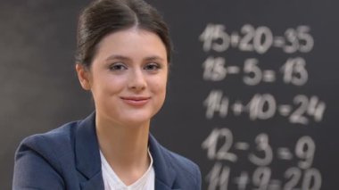 Genç öğretmen kameraya bakıyor, tahtada matematik denklemleri, yakın çekim