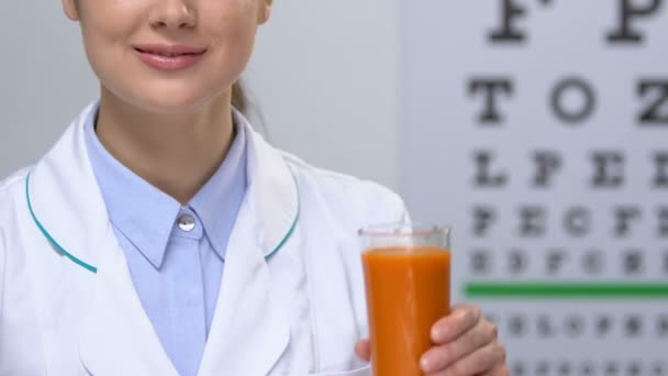 Улыбающаяся женщина-врач показывает морковный сок на фоне диаграммы глаза, здоровье — стоковое видео