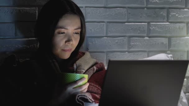Хвора жінка, покрита ковдрою, п'є гарячий чай вночі і працює на ноутбуці — стокове відео