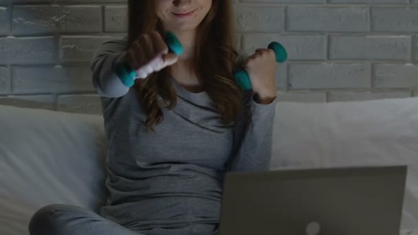 Nő képzés a súlyzók az ágyban, nézi fitness video-ra laptop, gyakorlása — Stock videók