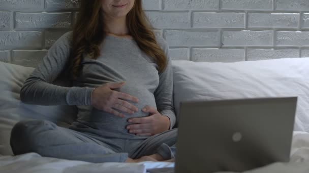 Mujer embarazada feliz acariciando la barriga, viendo lecciones de vídeo en el ordenador portátil por la noche — Vídeo de stock