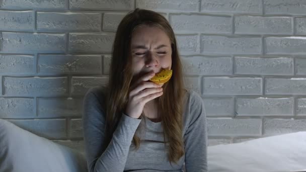 Плачущая женщина жует пончик ночью, стресс и проблемы с перееданием, булимия — стоковое видео