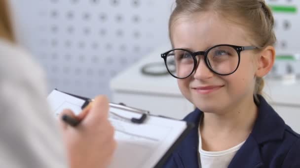 Escolar sonriente con anteojos mirando al oftalmólogo que escribe prescripción — Vídeo de stock