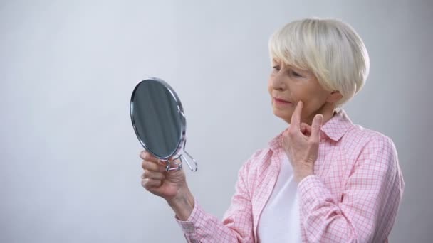 Donna anziana guardando la faccia rugosa nello specchio, pensando alla chirurgia plastica — Video Stock