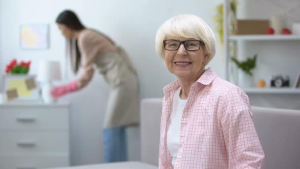 Sorrindo mulher idosa olhando para a câmera, governanta limpando poeira no quarto, limpeza — Vídeo de Stock