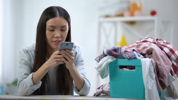 Улыбающаяся домохозяйка чатится на смартфоне возле корзины для белья с одеждой — стоковое видео