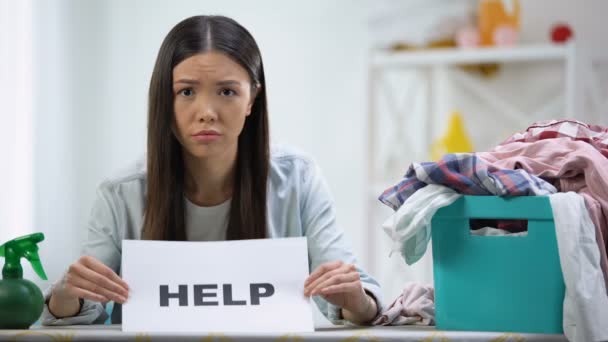 Mulher perturbada mostrando palavra de ajuda no cartão, cesta de roupa com roupas na mesa — Vídeo de Stock