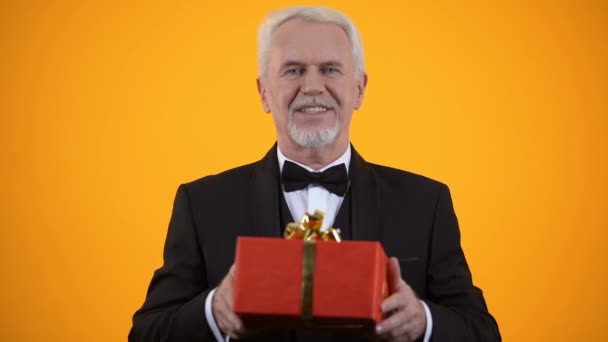 Веселый мужчина-пенсионер в костюме представляет красный подарочек на камеру, подарок на день рождения — стоковое видео