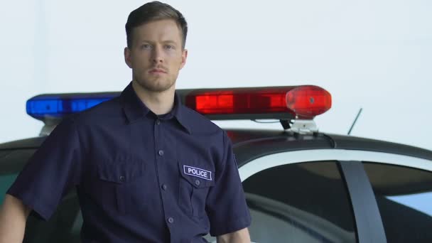 Pohledný policista stojící poblíž policajtového auta a dívá se do kamery, ve službě — Stock video