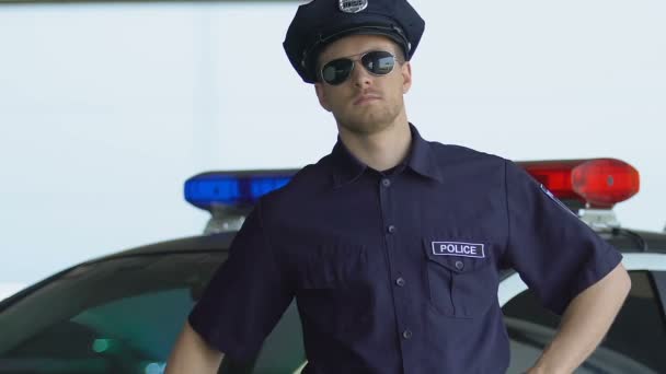 Професійний поліцейський в сонцезахисних окулярах стоїть біля автомобіля, районна охорона — стокове відео