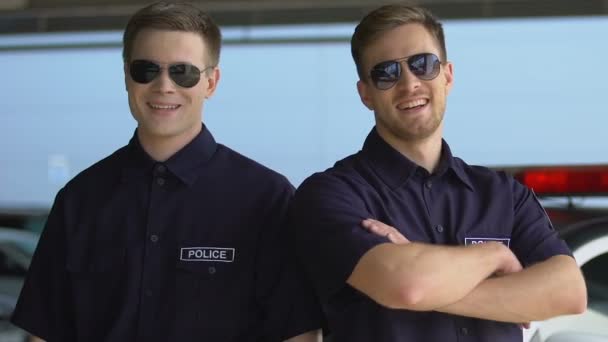 Beaux policiers en uniforme et lunettes de soleil debout contre une voiture de patrouille — Video