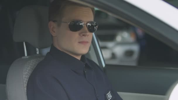 Ответственный полицейский в солнечных очках, сидящий в патрульной машине, улыбающийся в камеру — стоковое видео