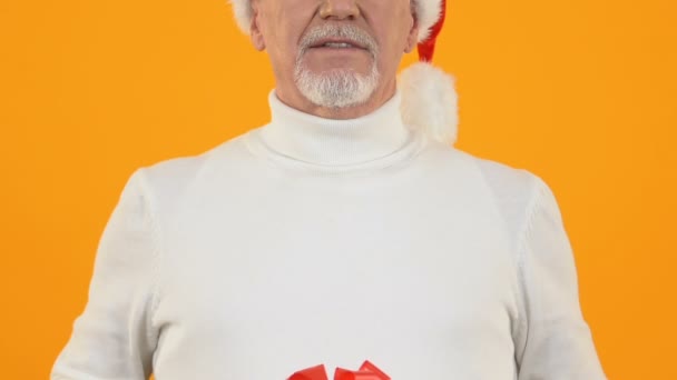 Счастливый зрелый мужчина в шляпе Санта показывая подарок с красной лентой, подарок сюрприз — стоковое видео