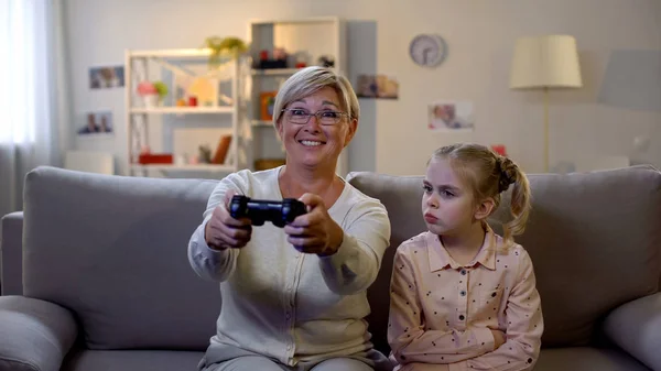 Возбужденная Бабушка Играет Видеоигры Игнорируя Грустную Внучку Сидящую Диване — стоковое фото