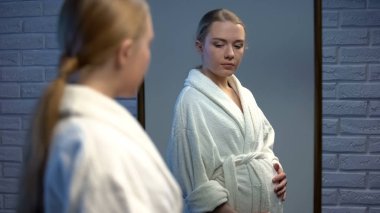 Teenage girl holding belly and looking in mirror reflection, early pregnancy clipart