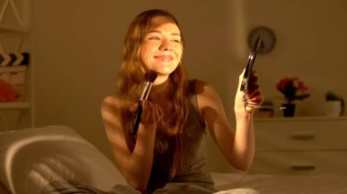 Mutlu genç bayan aynaya gülümseyerek ve yatakta allık tutarak uygulayarak, makyaj